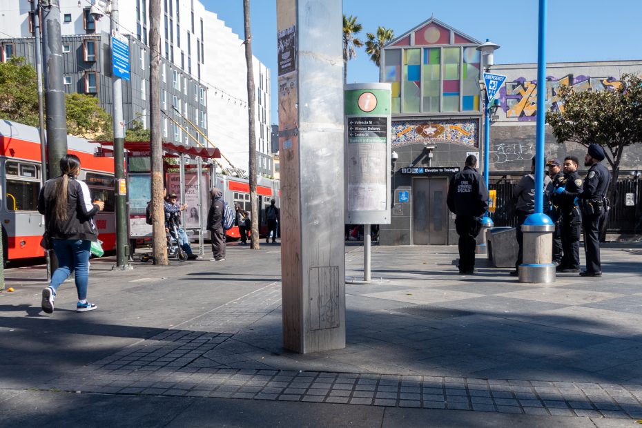 A city plaza with people walking, police officers standing in a group, buses on the left, and a colorful building in the background.
