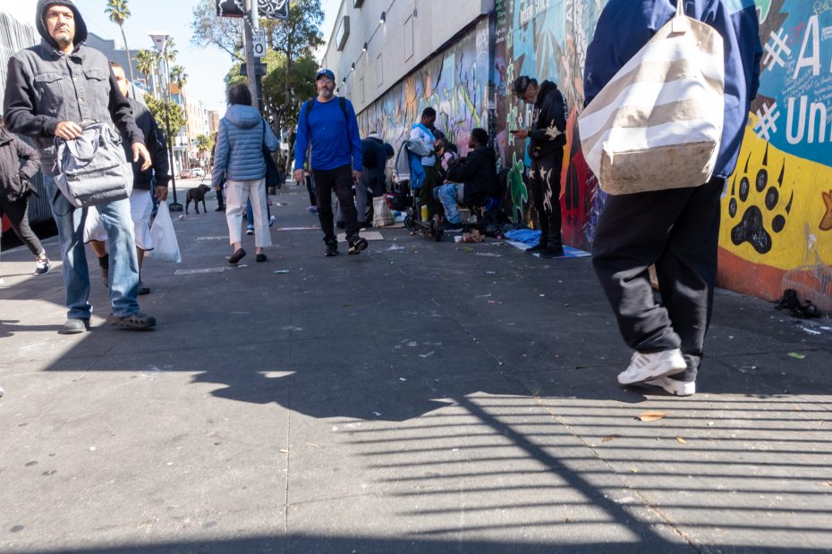 People walking and standing near a colorful mural on a city sidewalk; some individuals sit with belongings, suggesting homelessness or gathering.
