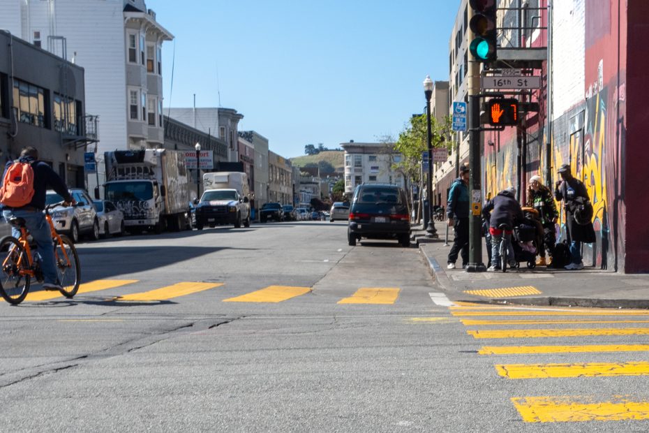 A city street intersection with cyclists, pedestrians waiting at a crosswalk, and buildings lining both sides under a clear sky.