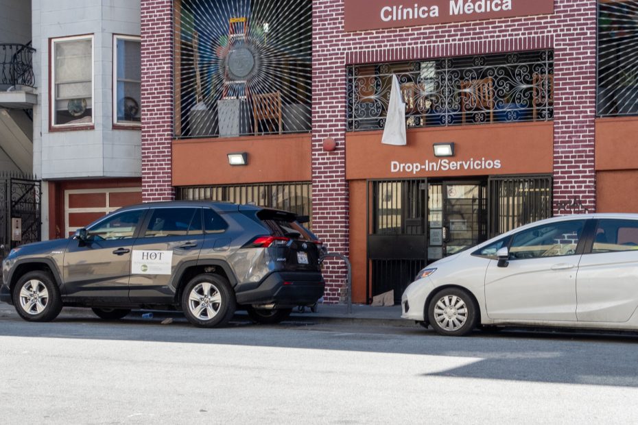 Two cars are parked on the street in front of a brick building labeled “Clínica Médica” with a “Drop-In/Servicios” sign above the entrance.