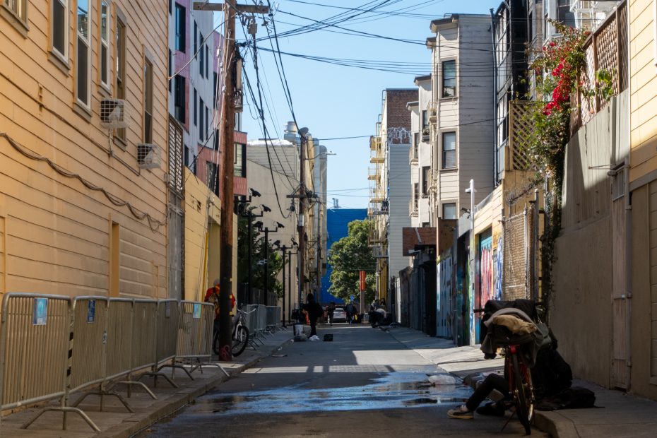A narrow city alley lined with buildings, barriers, and some people; a person sits on a bicycle near a puddle in the foreground.