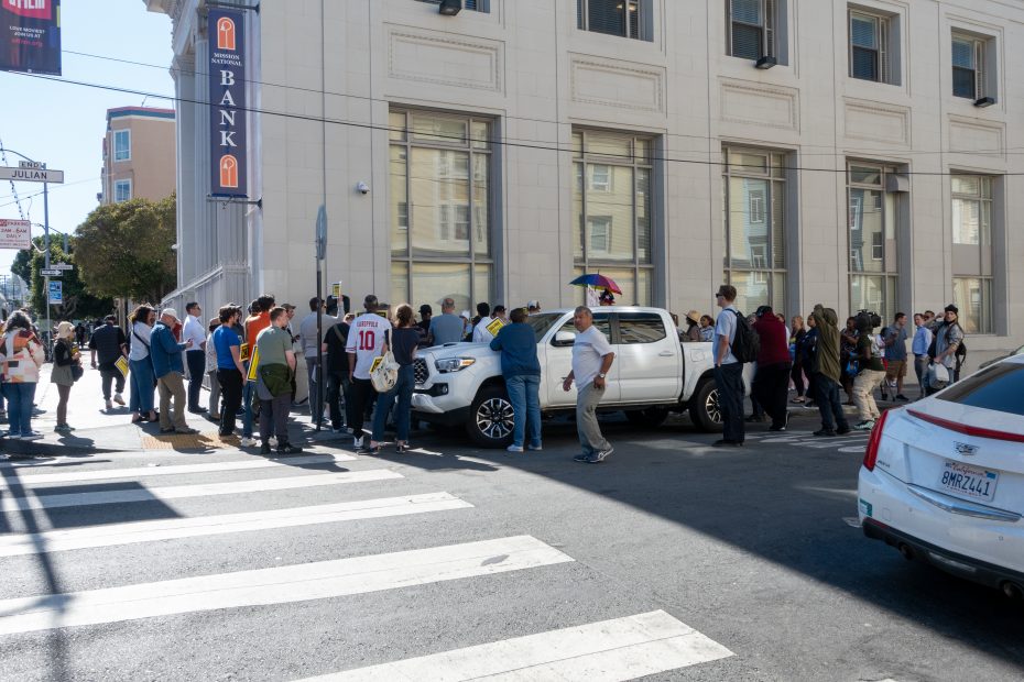 A crowd gathers on a street corner near a white truck and a bank building, with people standing on the sidewalk and street.