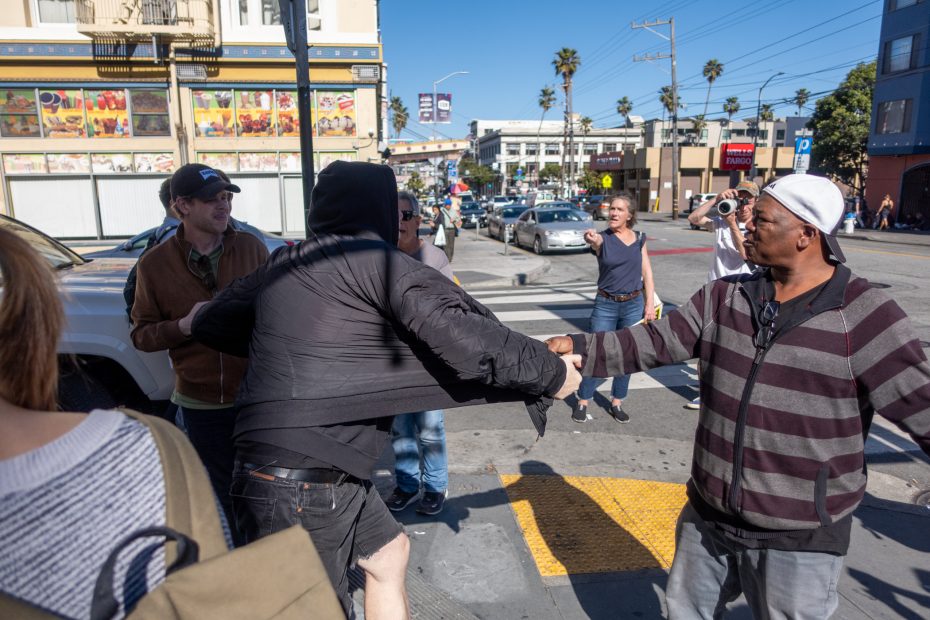 Two men shake hands on a busy city street corner while others stand nearby and a man in the background takes a photo.