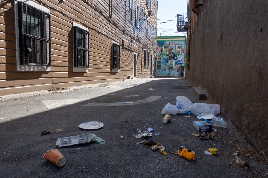 A narrow alleyway with litter scattered on the ground, bordered by buildings with barred windows; colorful mural visible at the far end.