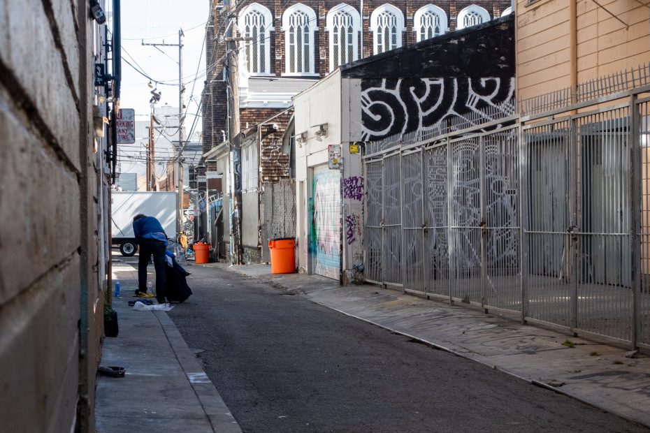 A person in a blue hoodie sorts through items on the ground in a narrow urban alley with graffiti, metal gates, and orange trash bins.