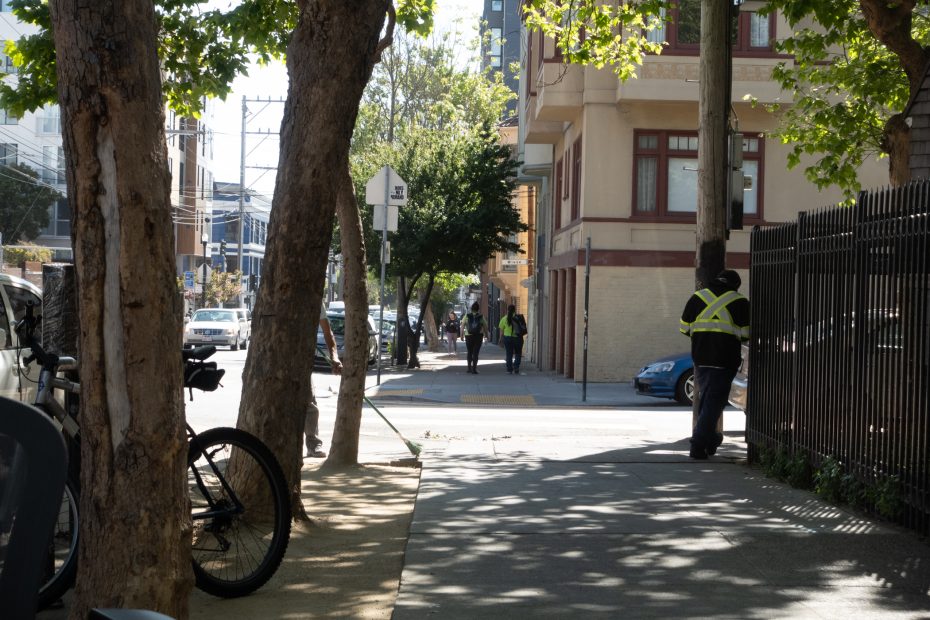 A person wearing a safety vest stands by a fence on a sunlit city sidewalk, with trees, parked cars, and buildings in the background.