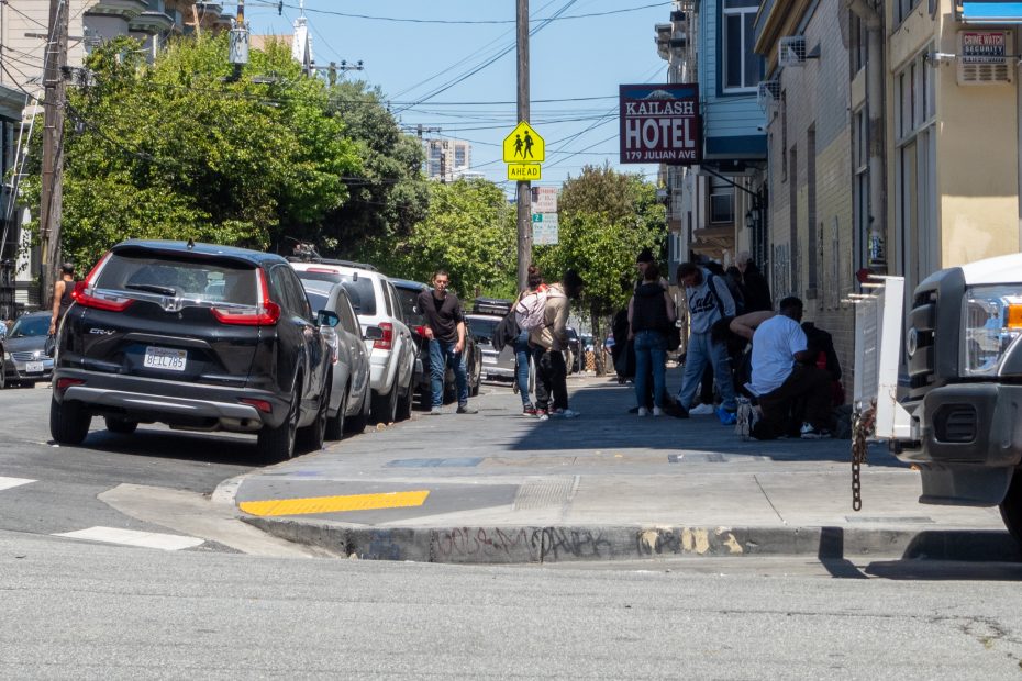 People are gathered on a city sidewalk near parked cars and a hotel sign on a sunny day. A pedestrian crossing sign is visible above.