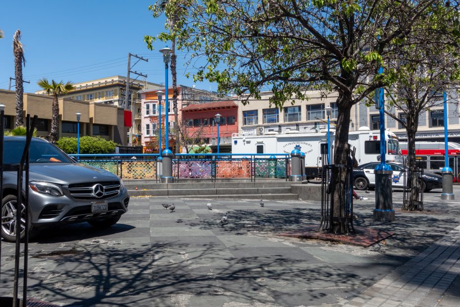 A city plaza with a parked silver Mercedes, pigeons on the ground, trees providing partial shade, and buildings in the background under a clear blue sky.