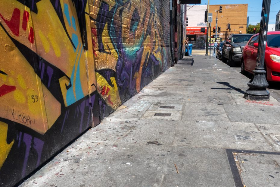 A city sidewalk with colorful graffiti on a brick wall, parked cars, and pedestrians in the background on a sunny day.