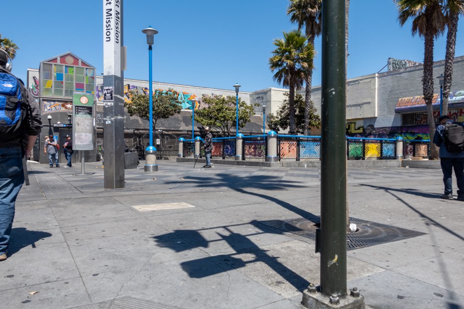 A sunny urban plaza with palm trees, murals, street lamps, and a few people walking near a transit station entrance.