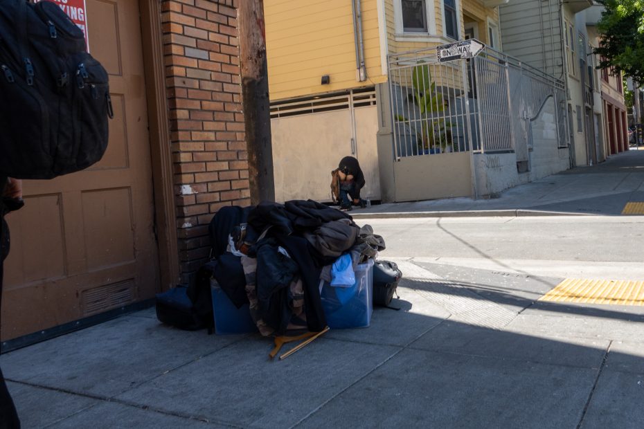 A person crouches on a sidewalk near a pile of belongings and clothing at the corner of a city street, with buildings and a street sign in the background.