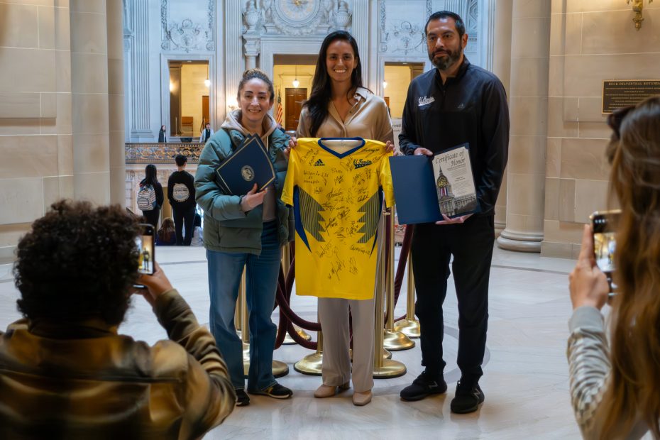 Three people stand indoors holding a signed yellow sports jersey and certificates while others photograph them in a hall with marble columns and ornate decor.