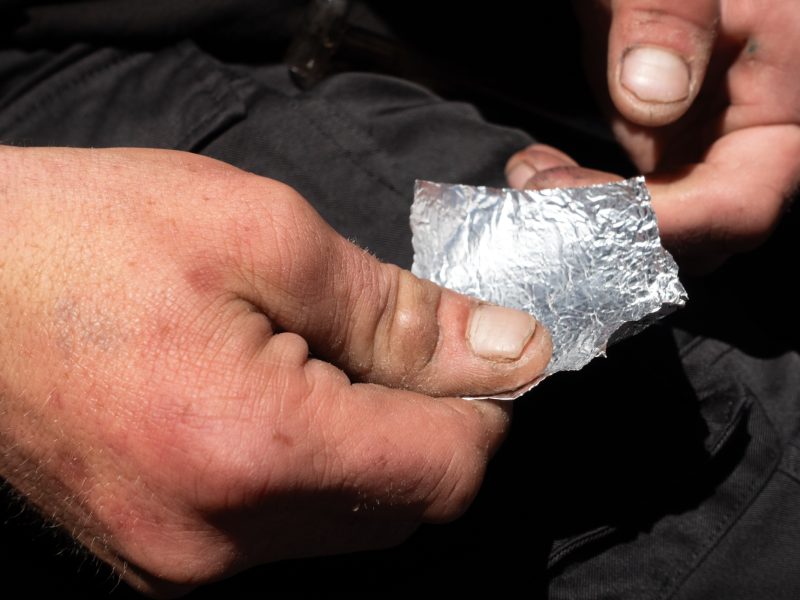 A close-up of two hands holding a small, crumpled piece of aluminum foil against a dark background.
