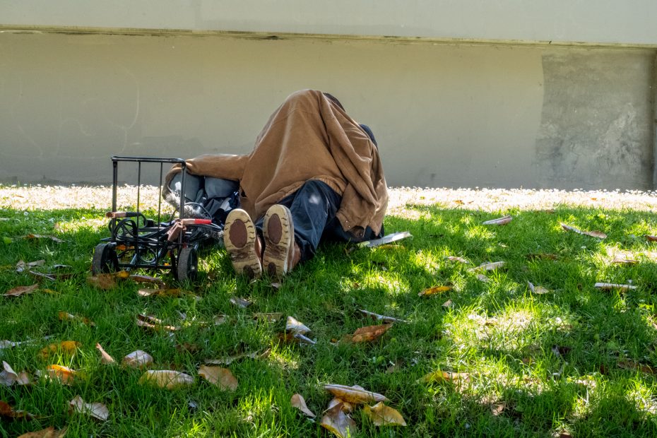 A person lies on the grass under a brown blanket, with their legs and shoes visible, next to a cart and scattered fallen leaves.