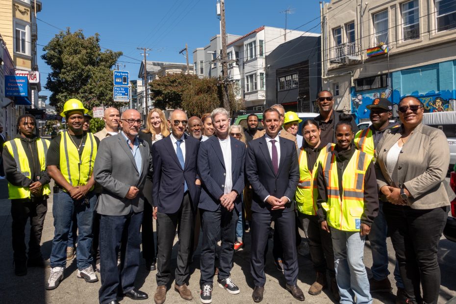 A group of people, including officials in suits and workers in safety vests and helmets, pose together on a city street lined with buildings.