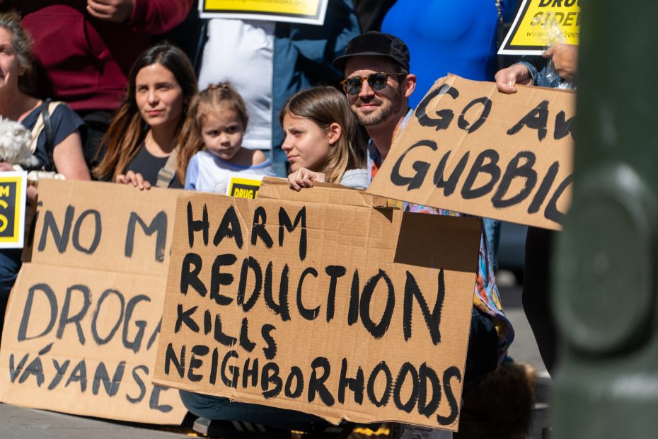 A group of people hold protest signs, including one that reads "Harm Reduction Kills Neighborhoods," during a demonstration.