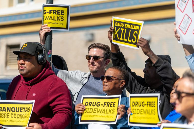 A group of people holding signs with messages such as “Drug-Free Sidewalks” and “Drug Enablism Kills” during a public demonstration.