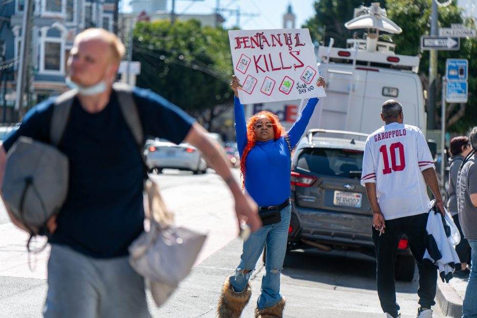 A person stands on a city sidewalk holding a “Fentanyl Kills” sign while others walk by. Cars and buildings are visible in the background.