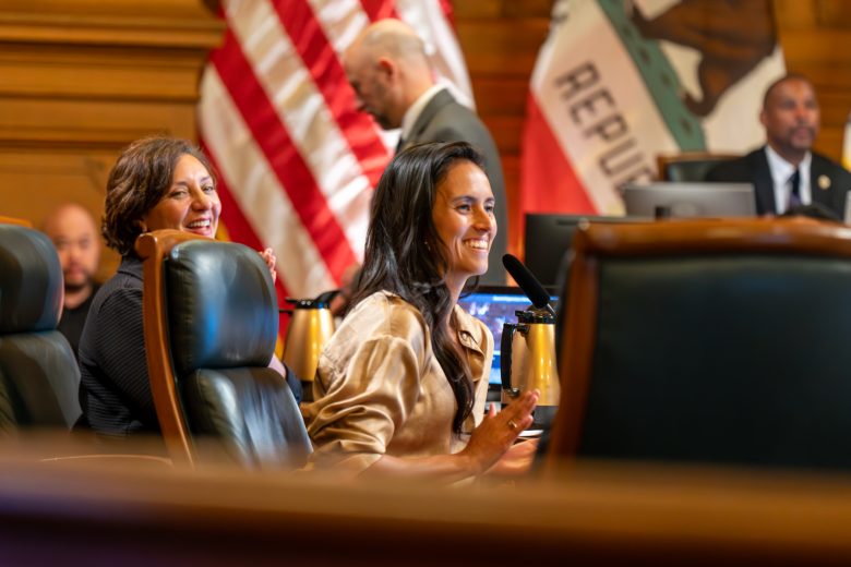 Two women seated at a council table smile and speak into microphones; American and California flags are visible in the background.