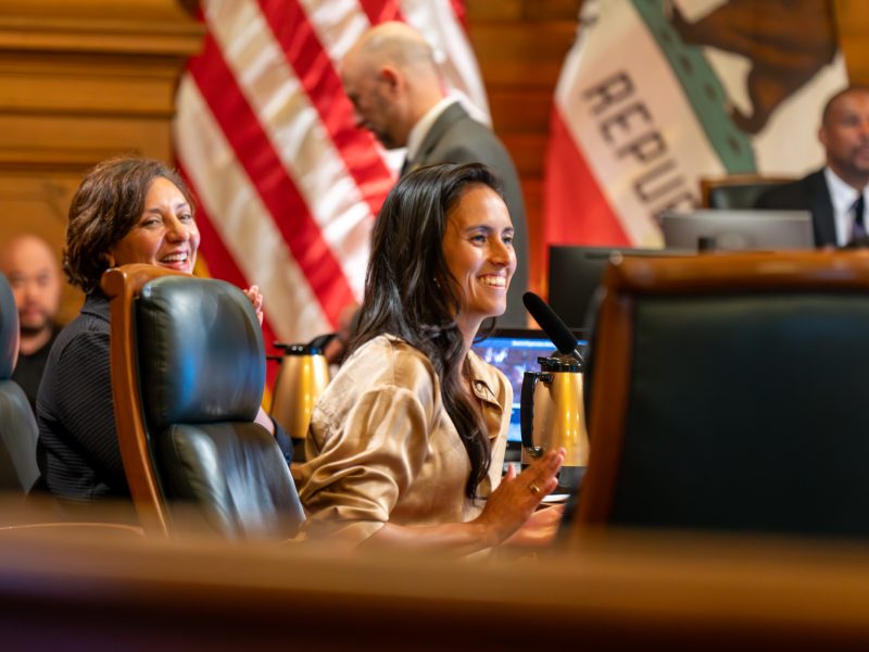 Two women seated at a council table smile and speak into microphones; American and California flags are visible in the background.