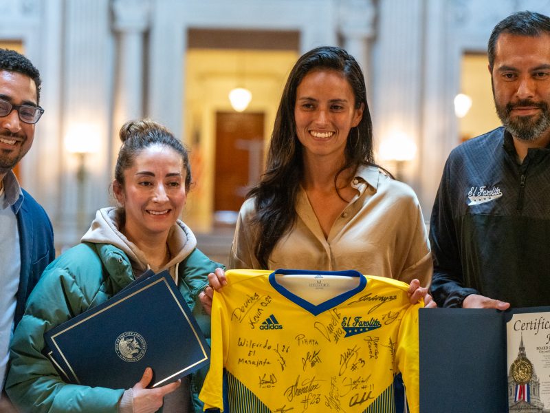 Four people stand indoors holding a signed yellow sports jersey, a certificate, and an official document, posing for a group photo in a formal setting.