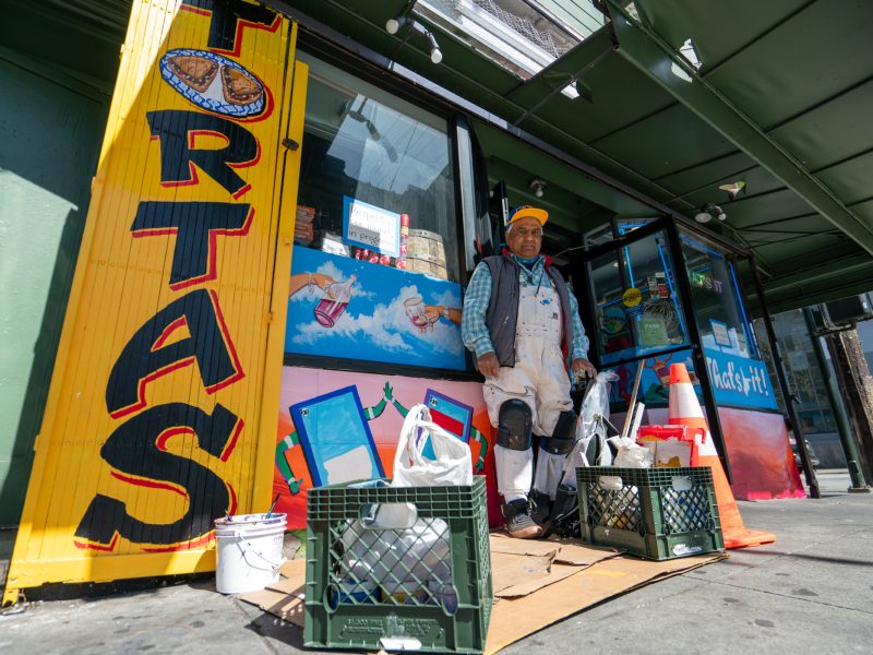A man in work clothes stands outside a storefront with a large "TORTAS" sign, next to crates holding supplies and bags on the sidewalk.