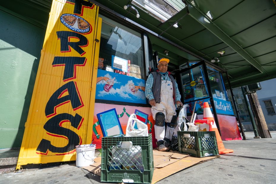A man in work clothes stands outside a storefront with a large "TORTAS" sign, next to crates holding supplies and bags on the sidewalk.