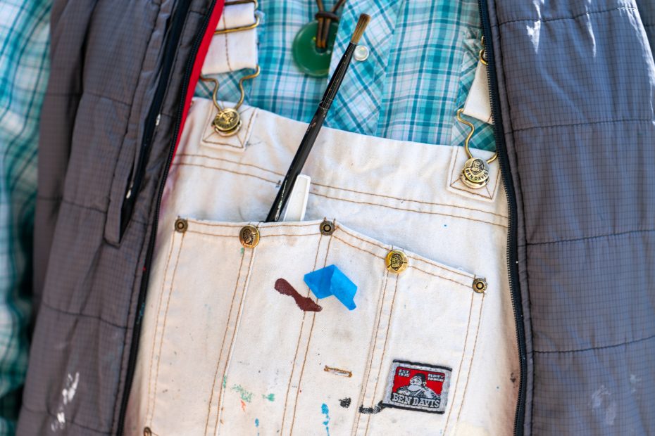 Close-up of a person wearing a paint-stained white apron over a plaid shirt and jacket, with brushes and a pen in the apron pocket.