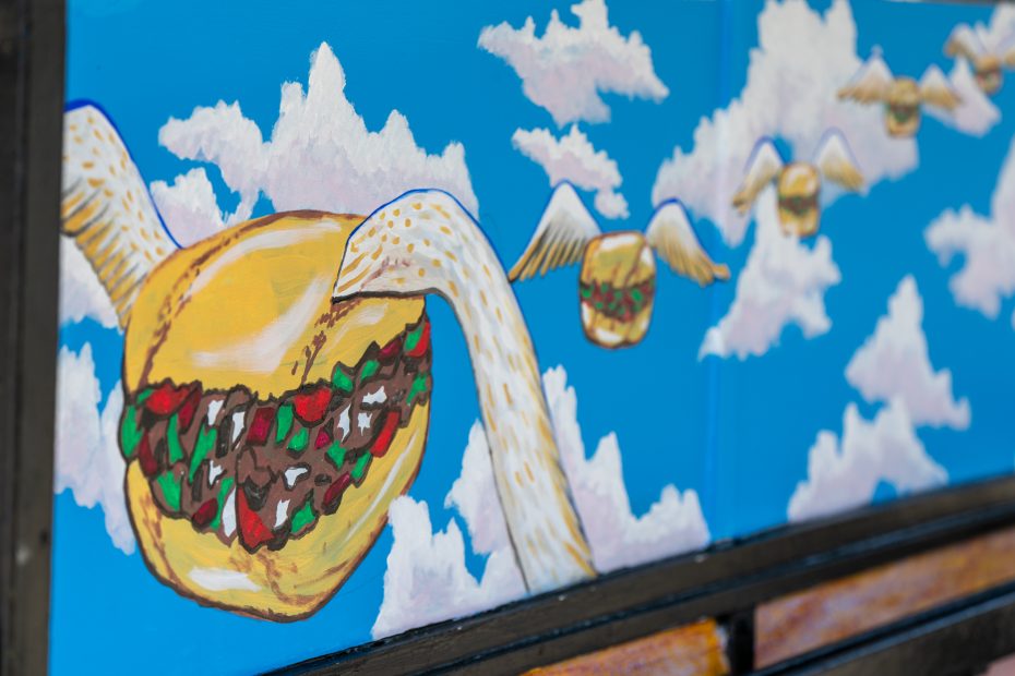 A mural depicts sandwiches with wings flying among clouds in a bright blue sky.