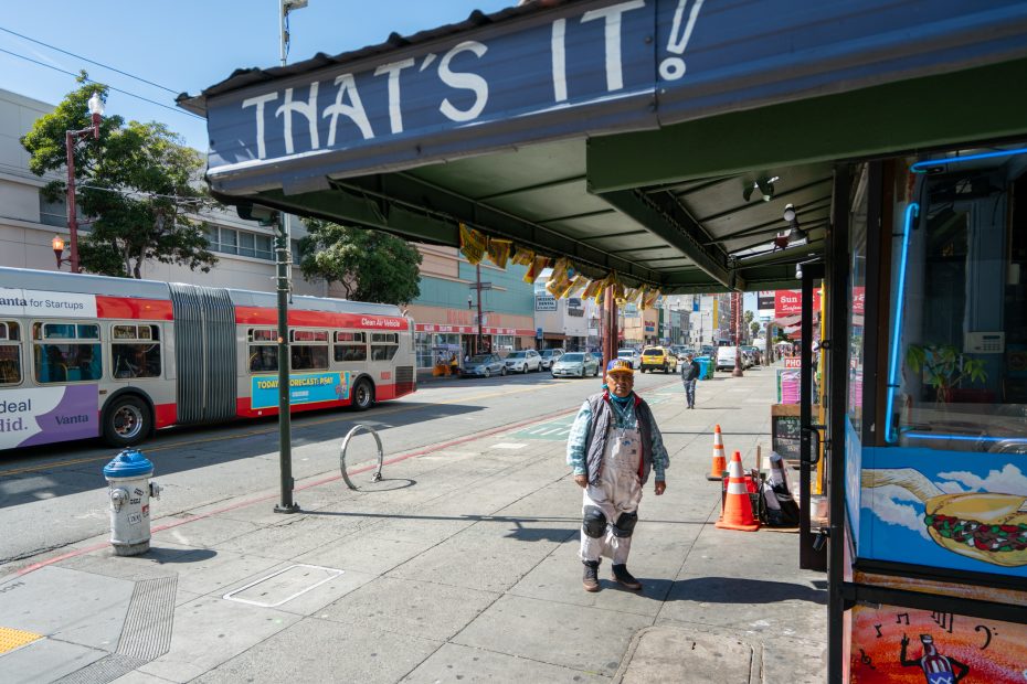 A person stands on a city sidewalk in front of a store with a sign reading "THAT'S IT!" Two buses and several parked cars are visible on the street.