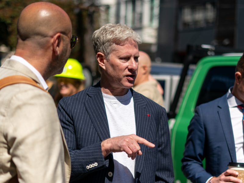 A man in a pinstripe blazer gestures while talking with another man in a beige jacket outdoors. Two other people, one in a suit and the other in a yellow hard hat, are in the background.