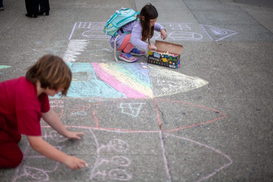 Two children draw colorful shapes and designs with chalk on a concrete surface; one uses a chalk box, and both have backpacks.