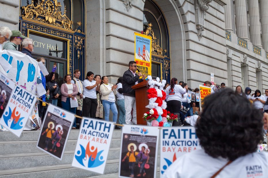 A speaker addresses a crowd on the steps of a government building during a Faith in Action Bay Area rally, with banners and signs displayed.