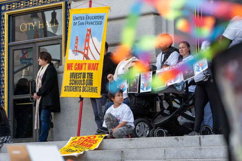 People gather on the steps of City Hall holding signs advocating for school investment, including a banner from United Educators of San Francisco. A child sits near the group.