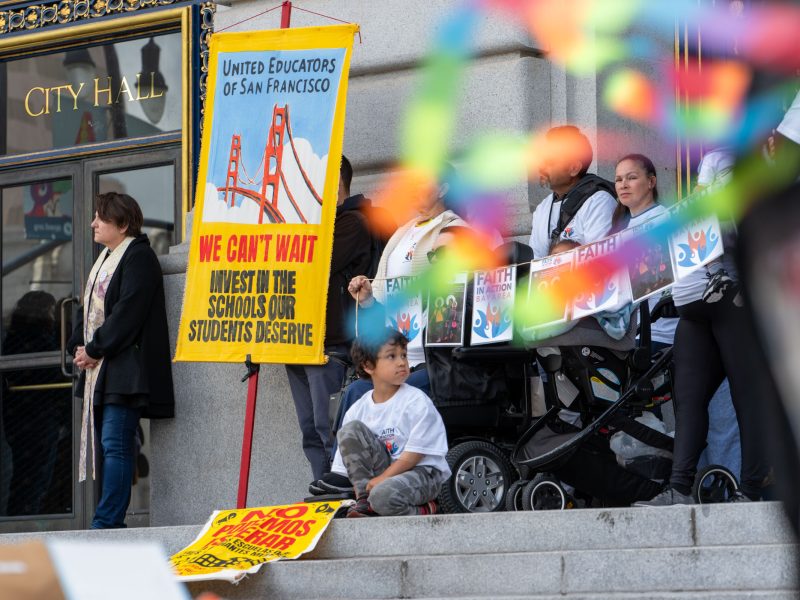 People gather on the steps of City Hall holding signs advocating for school investment, including a banner from United Educators of San Francisco. A child sits near the group.
