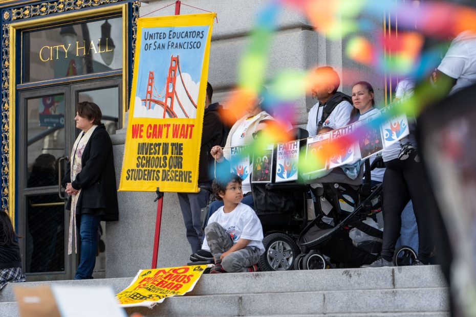 People gather on the steps of City Hall holding signs advocating for school investment, including a banner from United Educators of San Francisco. A child sits near the group.