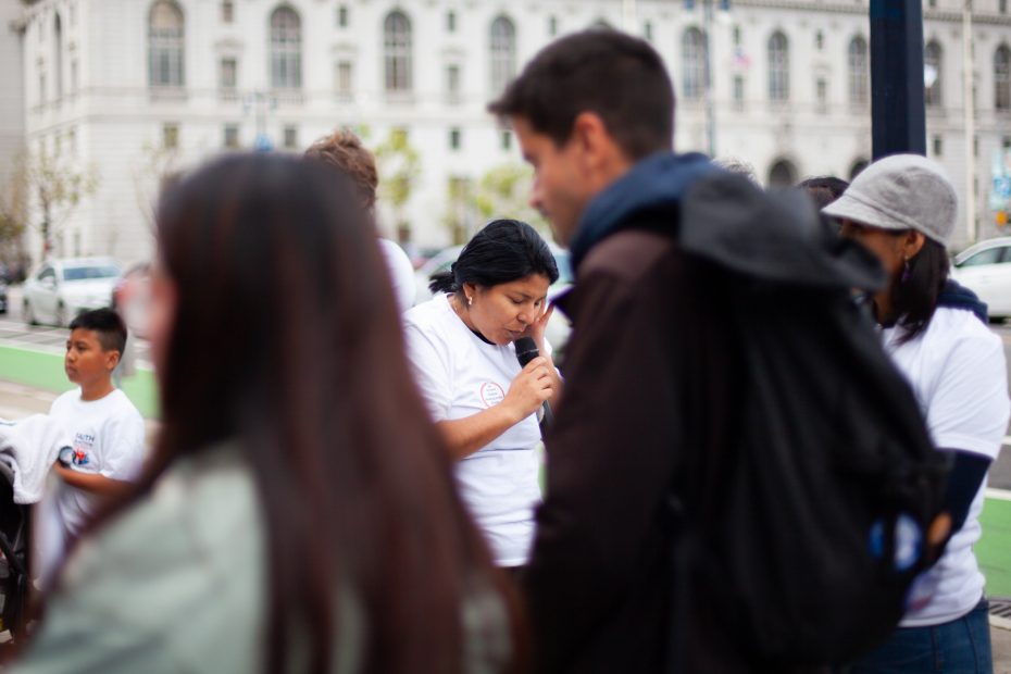 A woman speaks into a microphone at an outdoor gathering, surrounded by people, with a city building in the background.