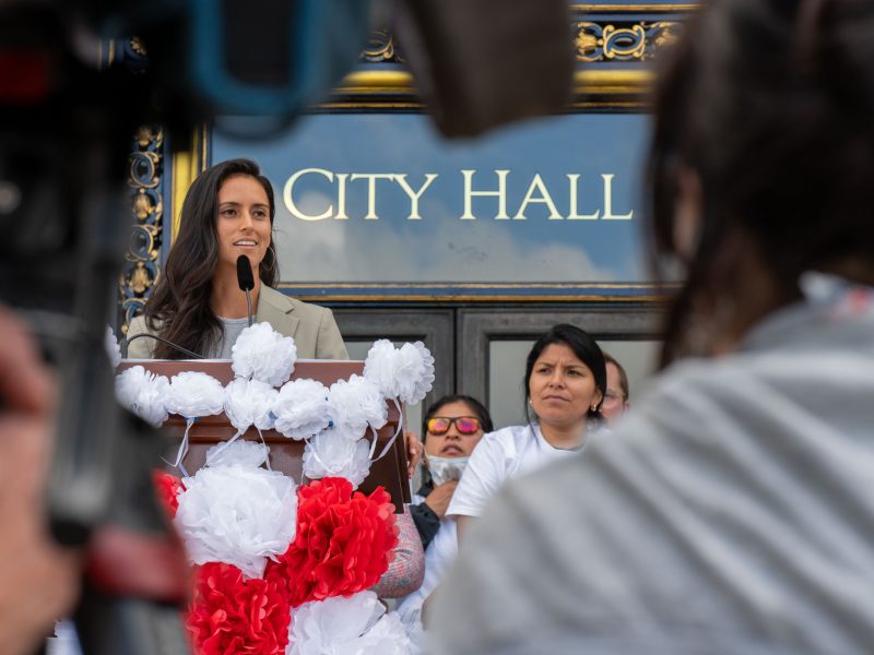 A woman speaks at a podium decorated with red and white flowers outside a building labeled "City Hall," with several people standing behind her.