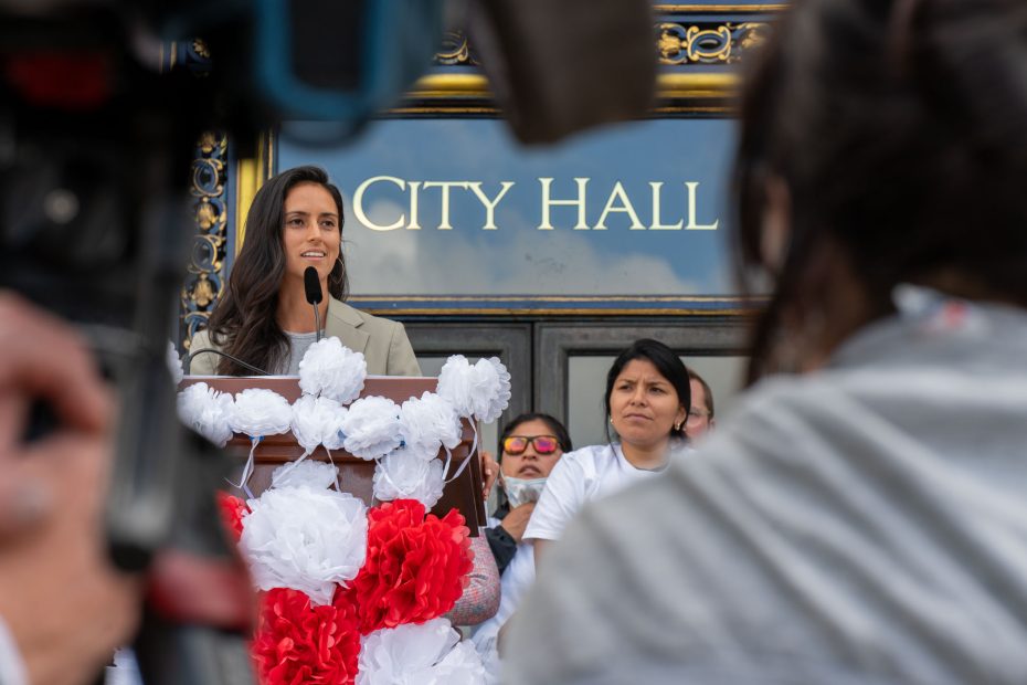 A woman speaks at a podium decorated with red and white flowers outside a building labeled "City Hall," with several people standing behind her.