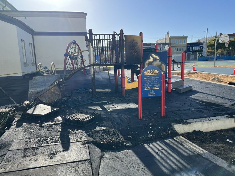 A playground structure with visible fire damage, including melted equipment, charred surfaces, and debris on the ground, adjacent to a welcoming sign and a school building.