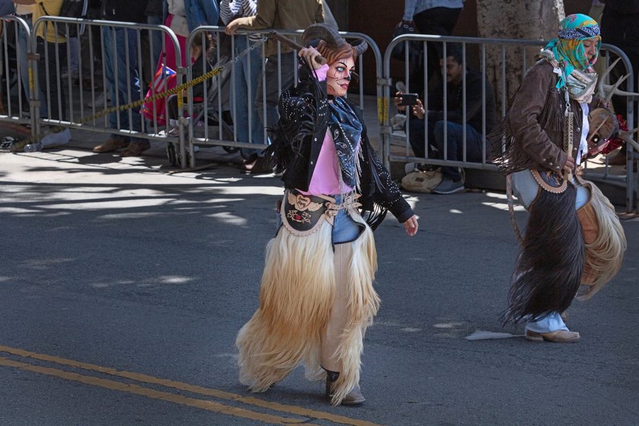 A person in a traditional costume with fur leggings, a painted face, and a decorative bag walks in a street parade beside another similarly dressed individual.