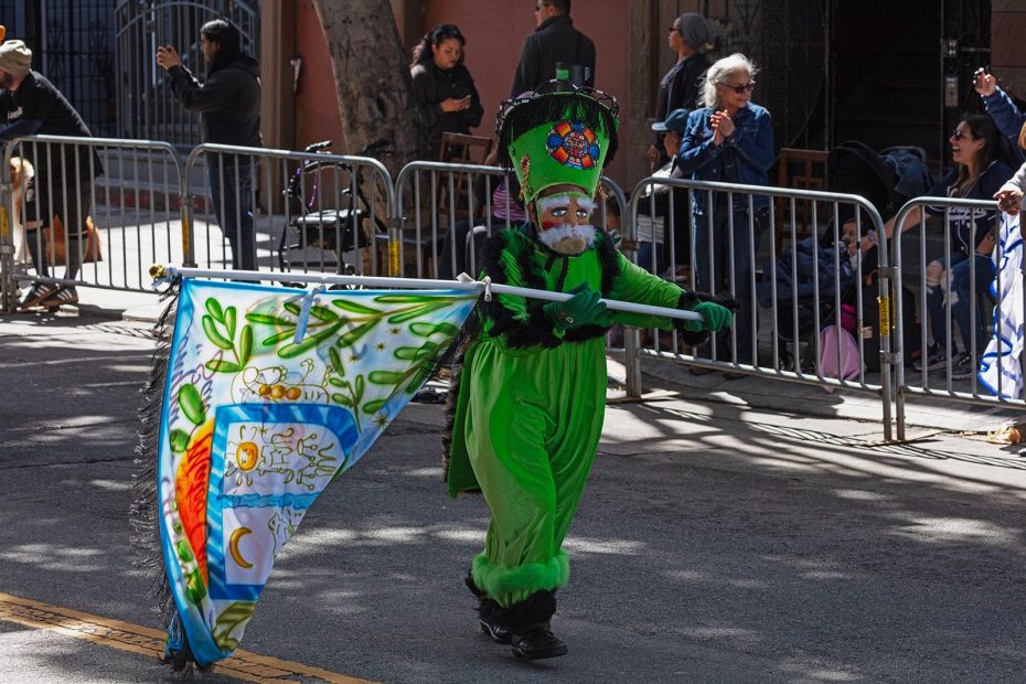 A person in a bright green costume and headdress carries a colorful flag while walking in a street parade, with spectators behind metal barricades.