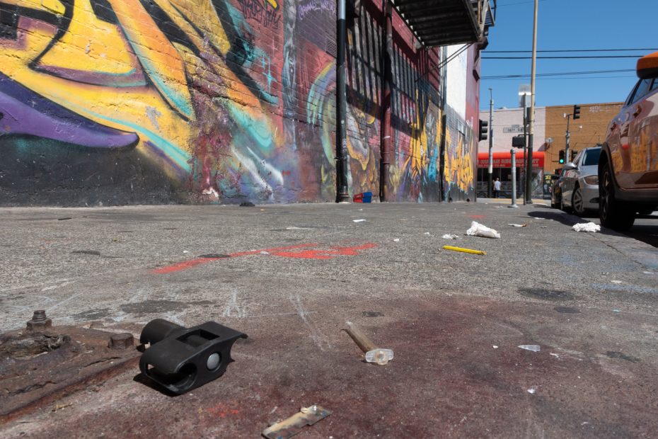 A littered urban sidewalk with various debris, colorful graffiti on the wall, parked cars, and a street scene in the background.