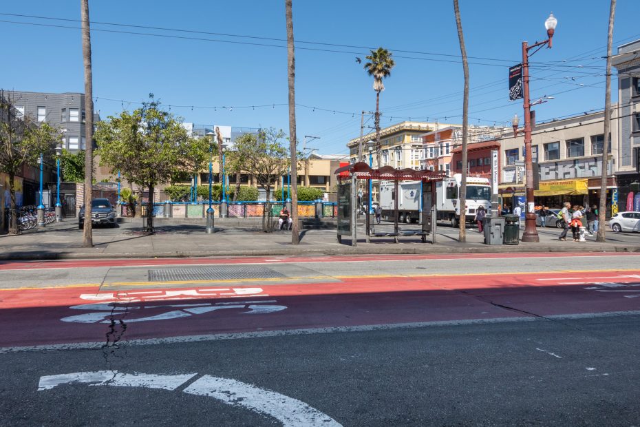 A city street with a red bus lane, a bus stop shelter, trees, and buildings in the background under a clear blue sky.