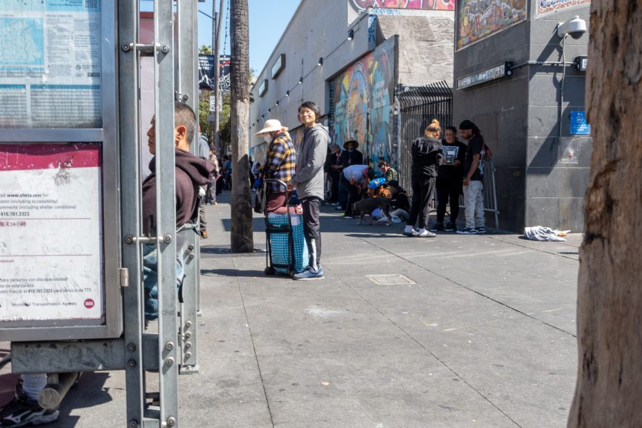 People stand and sit on a busy urban sidewalk near murals and a bus stop; some are gathered in small groups, with luggage and personal belongings visible.