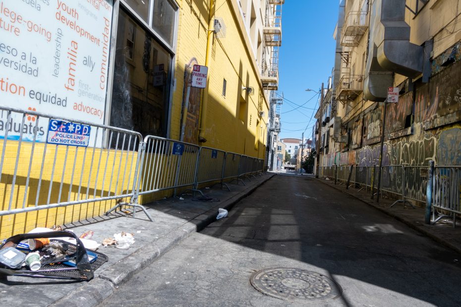 A narrow alleyway with yellow walls, metal barricades, scattered trash, and graffiti under clear blue sky. Signs and windows are visible along the buildings.