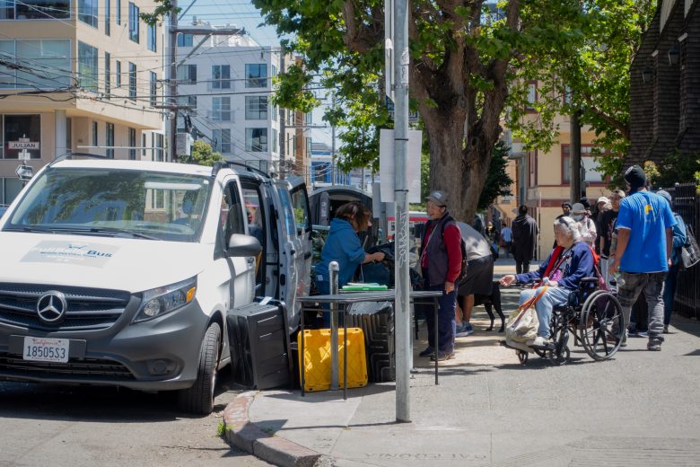People gather on a city sidewalk near a parked van, with one person in a wheelchair and others standing or sitting around tables with supplies.