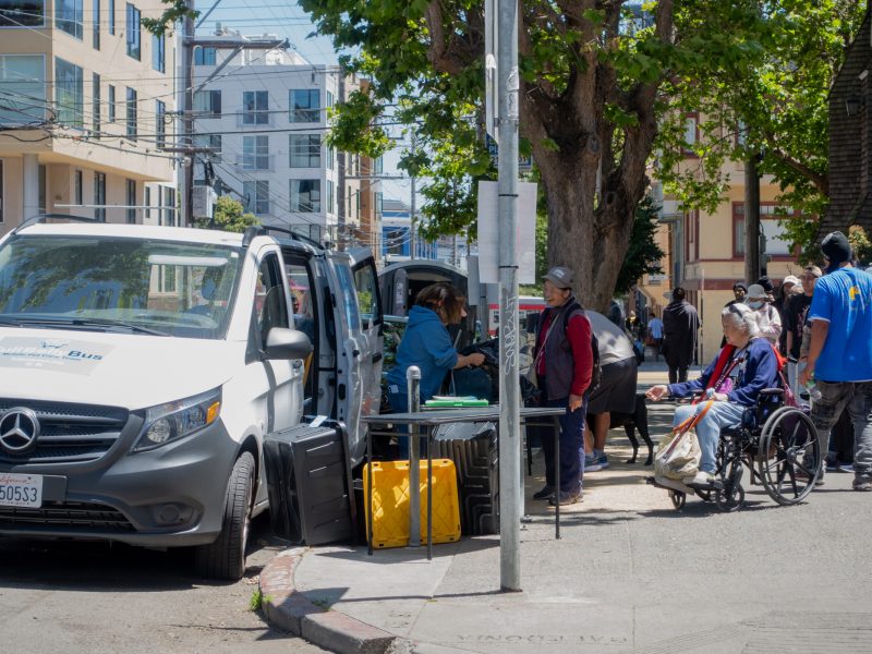 People gather on a city sidewalk near a parked van, with one person in a wheelchair and others standing or sitting around tables with supplies.