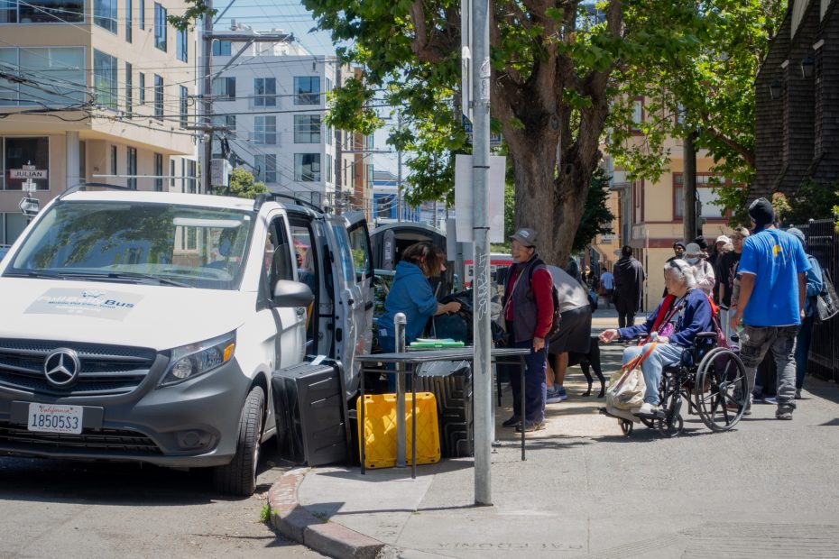 People gather on a city sidewalk near a parked van, with one person in a wheelchair and others standing or sitting around tables with supplies.