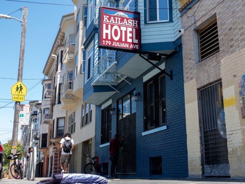 A person stands near the entrance of Kailash Hotel at 179 Julian Ave while another walks by on the sidewalk on a sunny day.